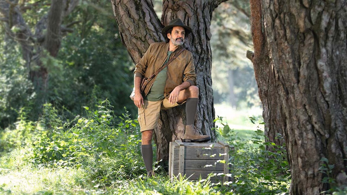Ein Mann mit Entdecker-Outfit steht in der Natur und schaut in die Ferne. Er trägt kniehohe Kompressionsstrümpfe.