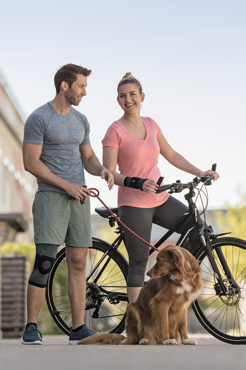 Eine Frau und ein Mann sind mit ihrem Hund unterwegs. Die Frau trägt eine Handgelenkbandage