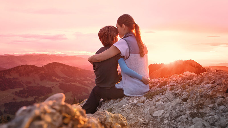 A mother is sitting on a mountain peak with her child. The child is wearing a compression sleeve.