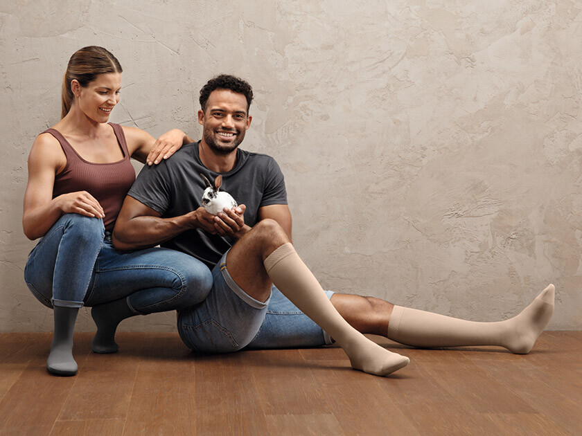 A man and a woman are sitting on the floor. Both are wearing compression stockings on their lower legs. The man is holding a small bunny in his hands