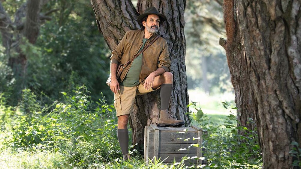 Ein Mann mit Entdecker-Outfit steht in der Natur und schaut in die Ferne. Er trägt kniehohe Kompressionsstrümpfe.