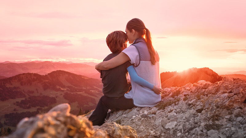 A mother is sitting on a mountain peak with her child. The child is wearing a compression sleeve.