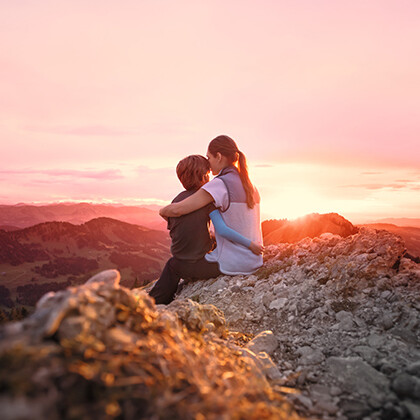 A mother sits on a mountain peak with her child in her arms and watches the sunset.