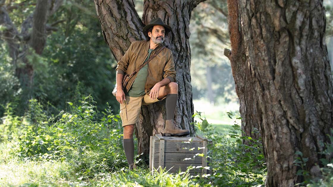 Ein Mann mit Entdecker-Outfit steht in der Natur und schaut in die Ferne. Er trägt kniehohe Kompressionsstrümpfe.