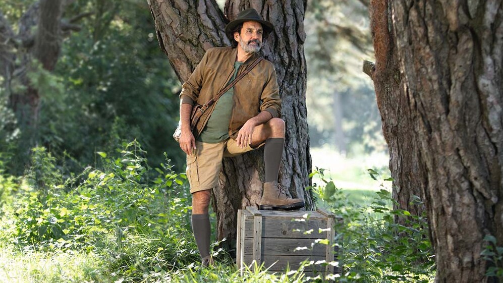 Ein Mann mit Entdecker-Outfit steht in der Natur und schaut in die Ferne. Er trägt kniehohe Kompressionsstrümpfe.