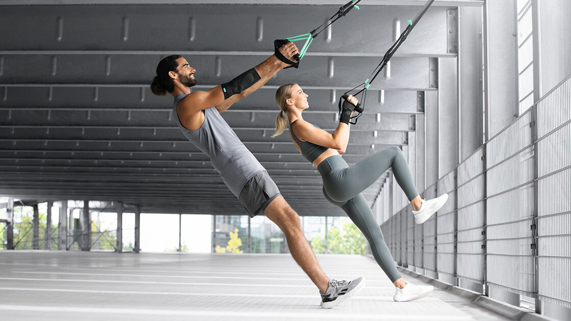 A man and a woman are working out in an underground car park using a TRX strap